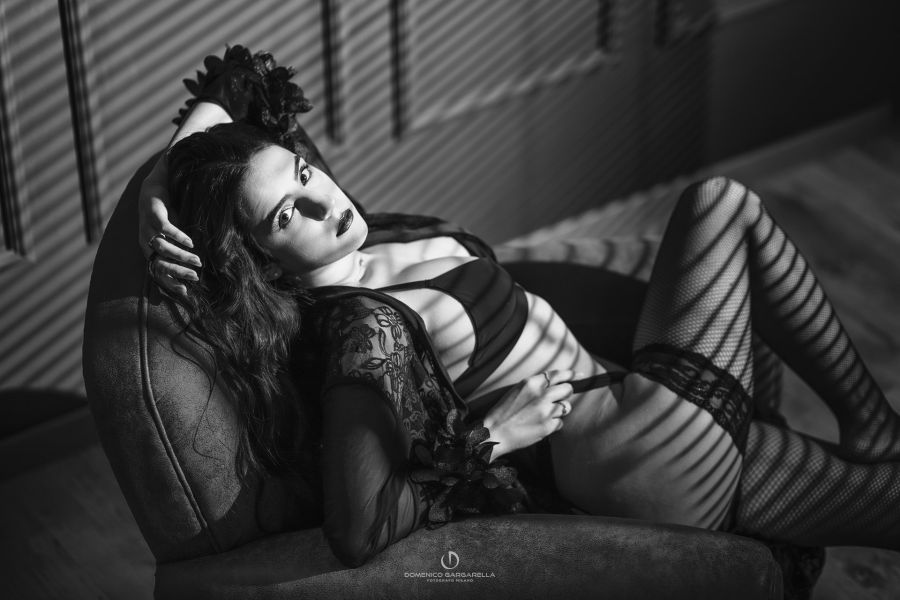 donna posa boudoir glamour seduta sdraiata su poltrona in lingerie nera e luce finestra con ombra tapparelle con espressione seducente