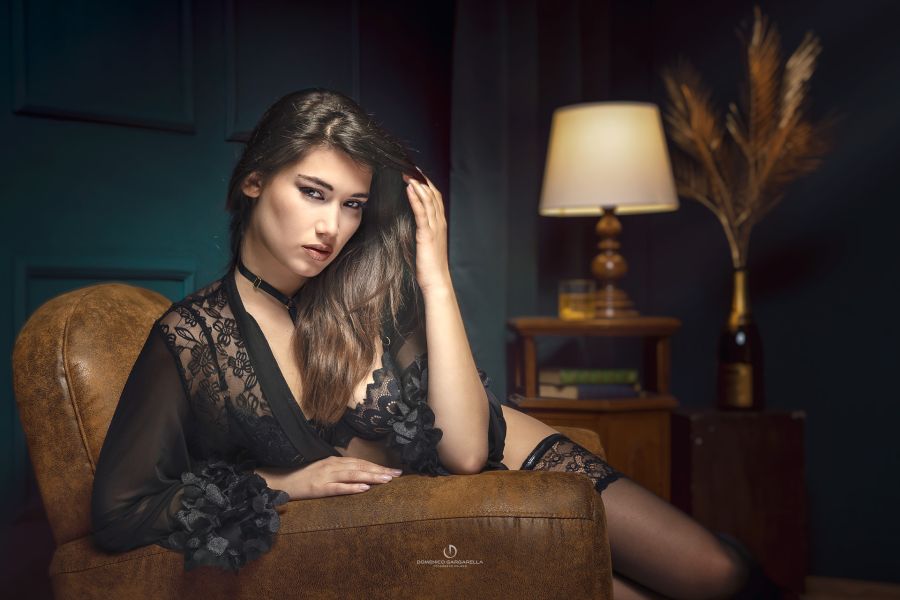 Fotografia Boudoir Glamour di ragazza seduta su poltrona pelle in linegerie nera e capelli lunghi neri